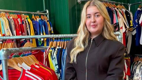 An 18-year-old girl with long blonde hair is looking directly at the camera as she's stood in her store surrounded by hundreds of old football shirts