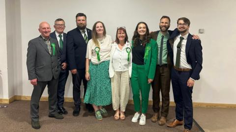 Eight people dressed in suits and formal clothes with green rosettes stand together. 