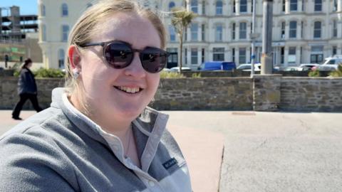 Melanie Harris has light brown hair tied up and has sunglasses on. She is wearing a grey fleece jumper and is smiling. She's standing on a concrete walkway with a stone wall behind her, with terraced buildings and parked cars beyond that, on a sunny day.