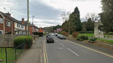 A Google street view of a single-carriage road in a residential area. Terraced houses are on the left of the road, while a park with a memorial is on the right. A number of cars are parked or driving on the road.