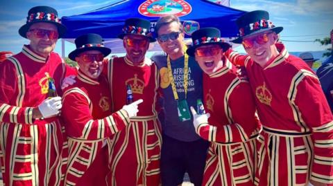 A group of men dressed up as British beefeaters stand in a line smiling, with a member of a cycling group. They wear black top hats with ribbons around the rim, red tunics with a crown across the chest, and sunglasses.
