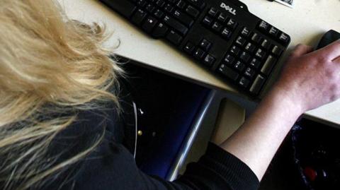 An image taken from above of an unidentified woman with long blonde hair operating a computer mouse at a keyboard