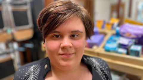 Head and shoulders image of Sydnii, who has short brunette hair swept to the right and is wearing a black top. Behind her, blurred out, are some essential products.