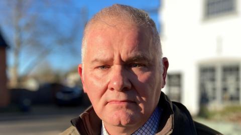 Independent, Councillor Clayton Hudson stood alongside a road. He has short grey hair, is cleanly shaven and is wearing a green coat over a shirt and tie.