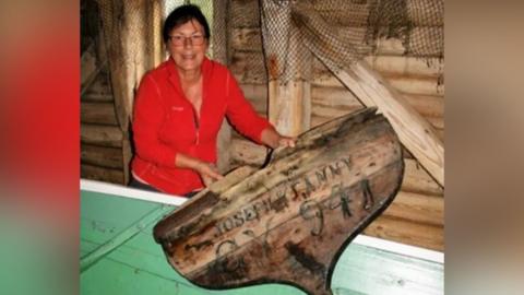 A woman with short dark hair smiles as she holds up an old piece of weathered brown wood from a ship's stern within a wooden cabin. She is wearing a red fleece and glasses. Marked on the wood are the words "Joseph & Fanny" and the registration number "GY-941".
