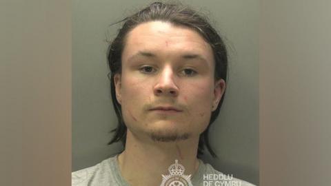 Leo Payne looks at the camera in this South Wales Police photo. He has shoulder length brown hair, pierced ears and some facial hair. He is wearing a grey T-shirt. Part of a South Wales Police logo can be seen.