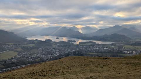 A view over Keswick taken from Latrigg Fell in the Lake District. The town sprawls from the foot of the fell, towards Derwentwater with more fells in the background. The sky is cloudy with rays of light shining through them.