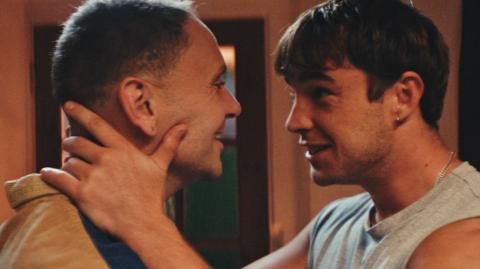 A side view of actors Lloyd Eyre Morgan (left) and Nico Mirallegro (right) in a sci-fi drama looking in each others eyes. X (left) has short brown hair wearing a beige jacket smiles at Mirallegro who has his hands on X's face who is also smiling.