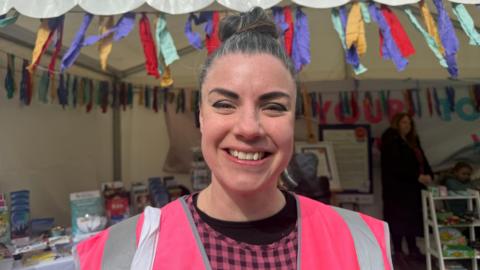 A woman with dark hair smiles into the camera. She is wearing a pink and black top with a pink and silver high-visibility jacket. The woman is standing in front of a market stall.