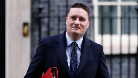 Health Secretary Wes Streeting, a white man with short brown hair, in a blue suit with blue tie, holding a red folder on Downing Street.