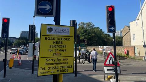 A large yellow road sign, in the centre of Bedford, showing a crossing with a number of traffic lights. A person is crossing the road, and cars are in the distance. There are lots of road signs in place. 
