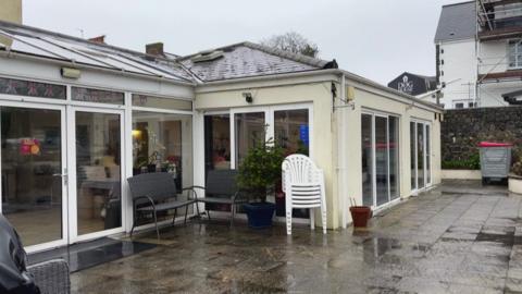 The exterior of Guernsey Cheshire Home. It is a white building with large windows. The paving stones are wet.