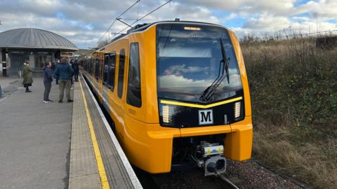 The first new Tyne and Wear Metro train in passenger service. The train is yellow and has the Metro M logo on its front. It is pulled into a station and passengers are waiting to board.