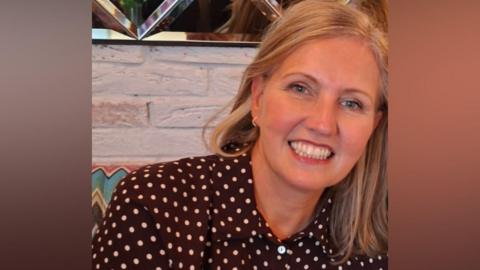 Joanne Mulholland is smiling brightly at the camera. She has long blonde hair and is wearing a brown shirt with polka dots on it. There is a mirrored wall behind her head.