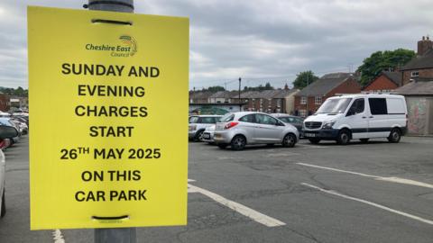 Car park with yellow sign topped Cheshire East Council reading: Sunday and evening charges start 26th May 2025 on this car park