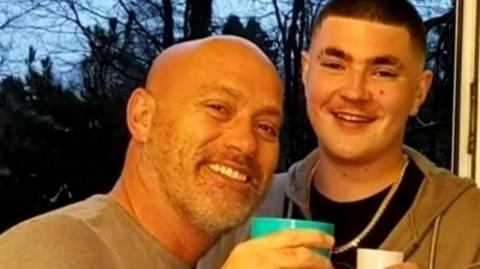 A selfie of two men smiling - the one on the left is bald and wearing a grey top while holding a green cup and the one on the right has short black hair, a chain around his neck, a black top and a grey hooded top. He is also holding a white cup. Trees can be seen in the background.