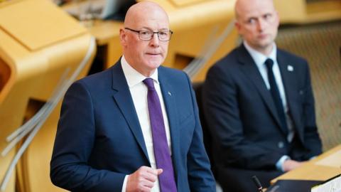 John Swinney, who is bald with glasses, in a business suit in the parliament chamber 