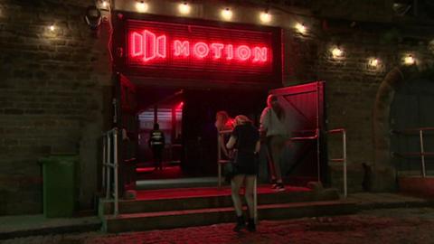 Women walking into the main entrance of Motion nightclub. It is dark and above the main entrance the Motion sign is lit up in red lights.