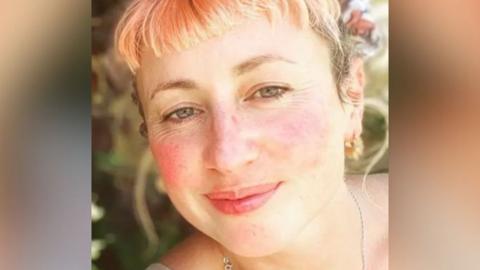 Tamsyn smiles at the camera. She has blonde and light pink hair, with a very short fringe, and is wearing gold earrings and a silver chain necklace. 