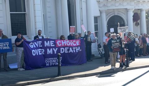 Campaigners outside Tynwald, some hold banners that read 'give me choice over my death', others say 'protect life'.