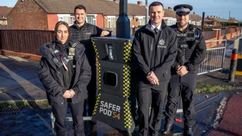 Four police officers are standing around the pod and are smiling at the camera. The pod is a tall yellow and black three-tiered structure with a camera at the top and a black screen in the middle. It stands no more than five feet tall but is attached to a larger pole with four horizontal legs at the bottom. Words on the device read: "Safer Pod S4". Behind the officers is a row of bungalows, with more houses stretching into the distance.