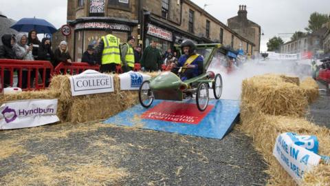 A man wearing a dark helmet and blue overalls smiles as he steers a green Thunderbirds-style cart down a ramp.