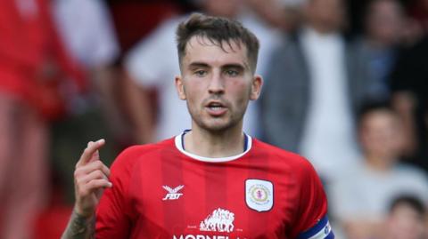 Luke Offord in action for Crewe wearing a red home shirt with the blue and white captain's armband on his left arm