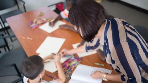 A woman wearing a blue and white shirt leans over a desk where two children are completing work.