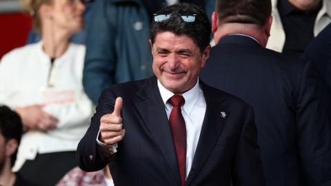 Southampton owner Dragan Solak, dressed in a suit with a maroon tie and wearing sunglasses on the top of his head, gives a thumbs-up gesture at a football game