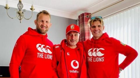 Three men are wearing red Lions rugby clothes. Scott, on the right, is a man in his 40s with blonde hair and sunglasses on his head.