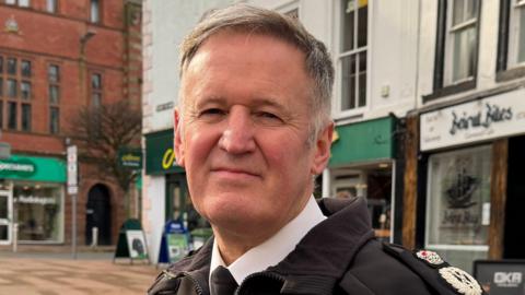 Darren Martland is standing in Carlisle city centre and looking directly at the camera. He has a neutral expression and has short brown/grey hiat. He is wearing a black police jacket, with a blue design on the front, over a white shirt and black tie. His epaulettes are embroidered with a silver and red design.