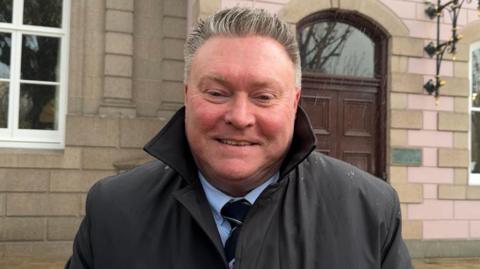 A man wearing a black coat with the collar up standing outside a government building in the rain.