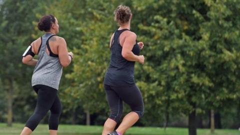Two women jog through a leafy green park. Both wear vest tops and leggings. One of the women, on the left, also wears a running armband to hold her phone.