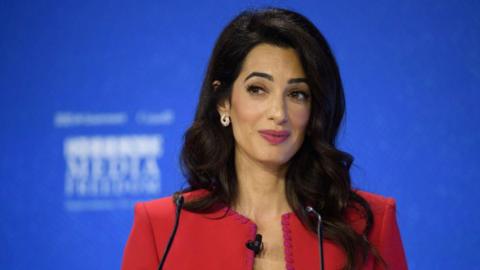 Amal Clooney wears a smart red jacket, and stands at a podium, possibly giving a speech. She has long dark hair which comes past her shoulders.