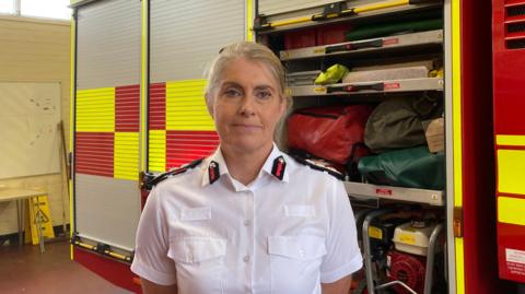 Nikki Watson, chief fire officer, in a white buttoned shirt with epaulettes on the shoulder and collar. She is standing inside a fire station with a red yellow and grey fire engine parked behind her and partly visible.