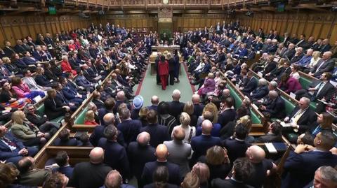 The chamber of the House of Commons in Westminster, London, crowded with Members of Parliament seated and standing around a central aisle. They are gathered to hear the result of the vote on the Terminally Ill Adults (End of Life) Bill. Green benches line both sides, and the Speaker's chair is visible at the far end beneath a clock