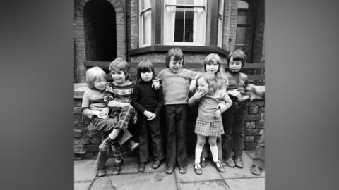 Black and white photograph of seven young children aged three to seven, in typical 1970s clothing, stood on a residential street with a Victorian house behind