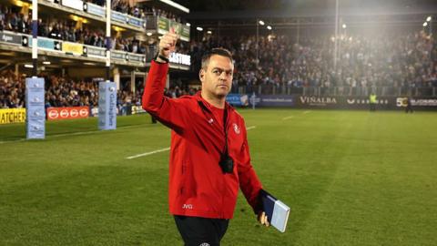 Johann van Graan holds his arm in the air and gives a thumbs up to the crowd while walking on the pitch at the Recreation Ground