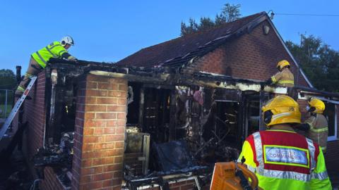 Firefighters work at the scene of a blackened, burnt-out bungalow. A room is exposed and the insides are charred. One of the firefighters is on a ladder.
