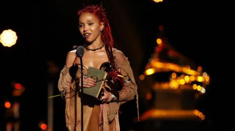 FKA Twigs, who has long, red hair and is wearing a brown outfit, accepts her Grammy on stage as she holds onto the award and a note book.