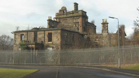 A dilapidated school building behind a metal fence - parts of the roof is missing. 