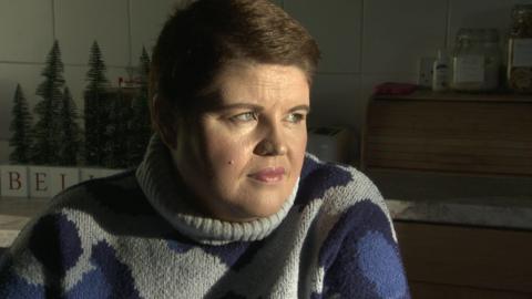 Emma - a woman with short brown hair has her head turned from the camera looking out the window as sun lights up her face. She is sitting down in a kitchen and is wearing a grey turtleneck jumper with large blue spots.