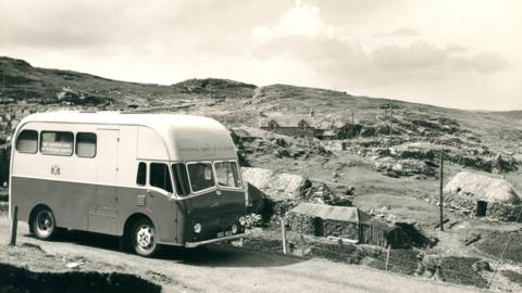 Dealbh dubh is geal de bhana Banca Rìoghail na h-Alba ann am baile beag Gàidhealach. Tha taighean-tughaidh ann cho math ri taigh-croite nas ùire. 