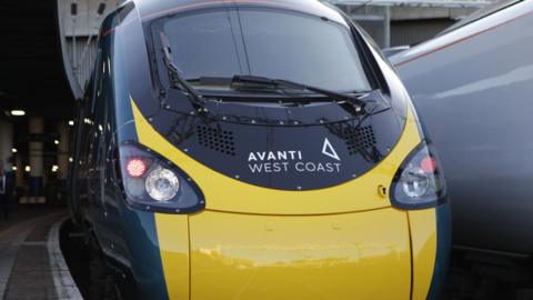 An Avanti West Coast Train. It has the branding of the train company on the front, and is a dark blue and yellow colour. The train is stationary at a railway station.