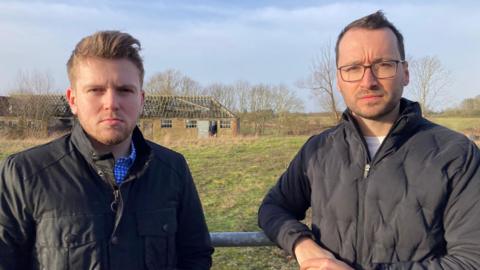 Two men stand in front of a field with a derelict building in the background. They both have short dark hair and are wearing dark jackets.