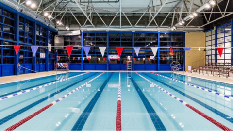 An swimming pool with nobody in it, with red, white and blue lane dividers in place. There is bunting of the same colours hanging in two lines above the pool and seating for spectators visible on the right hand side of the picture.