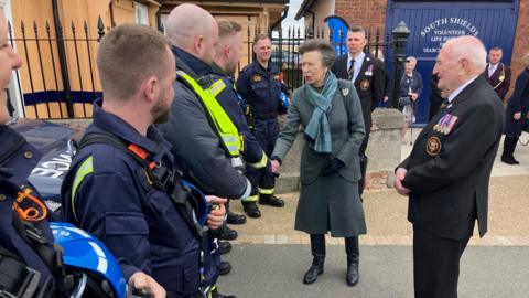The Princess Royal with her hair tied back. She has a long grey coat, a blue scarf, black gloves, black trousers and black boots. She is shaking hands with members of the South Shields Volunteer Life Brigade in their blue uniforms. There are also dignitaries in uniform. There is a brick building in the background with blue doors.