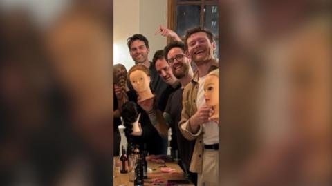 Four men smiling at the camera and gesturing to plastic heads with plaited hair.