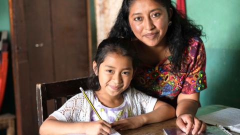 A woman stands next to a seated child holding a pencil