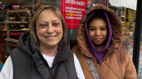 
Image shows two women one in a white top with a sleeveless padded jacket.
The other younger woman is wearing a brown padded jacket with the hood up.